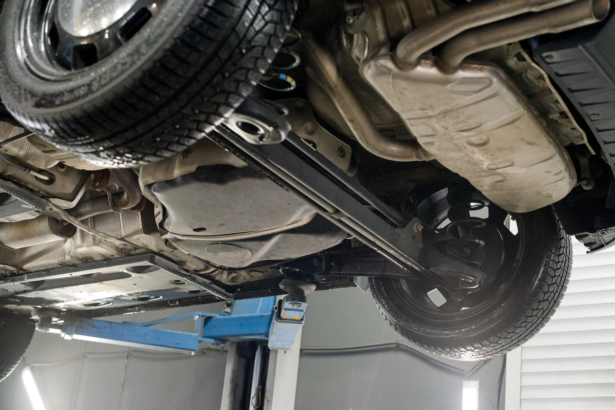 Modern car lifted on a hydraulic platform in an auto repair shop showing its undercarriage, exhaust system, and rear suspension while a technician performs maintenance work underneath